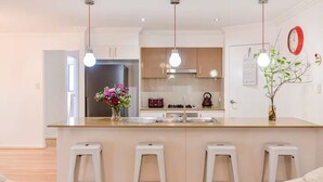 A modern kitchen with a central island, white bar stools, pendant lights, and a visible refrigerator.