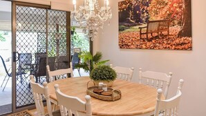 A dining area with a round table, white chairs, a chandelier, a potted plant, and a painting of a bench in a park.