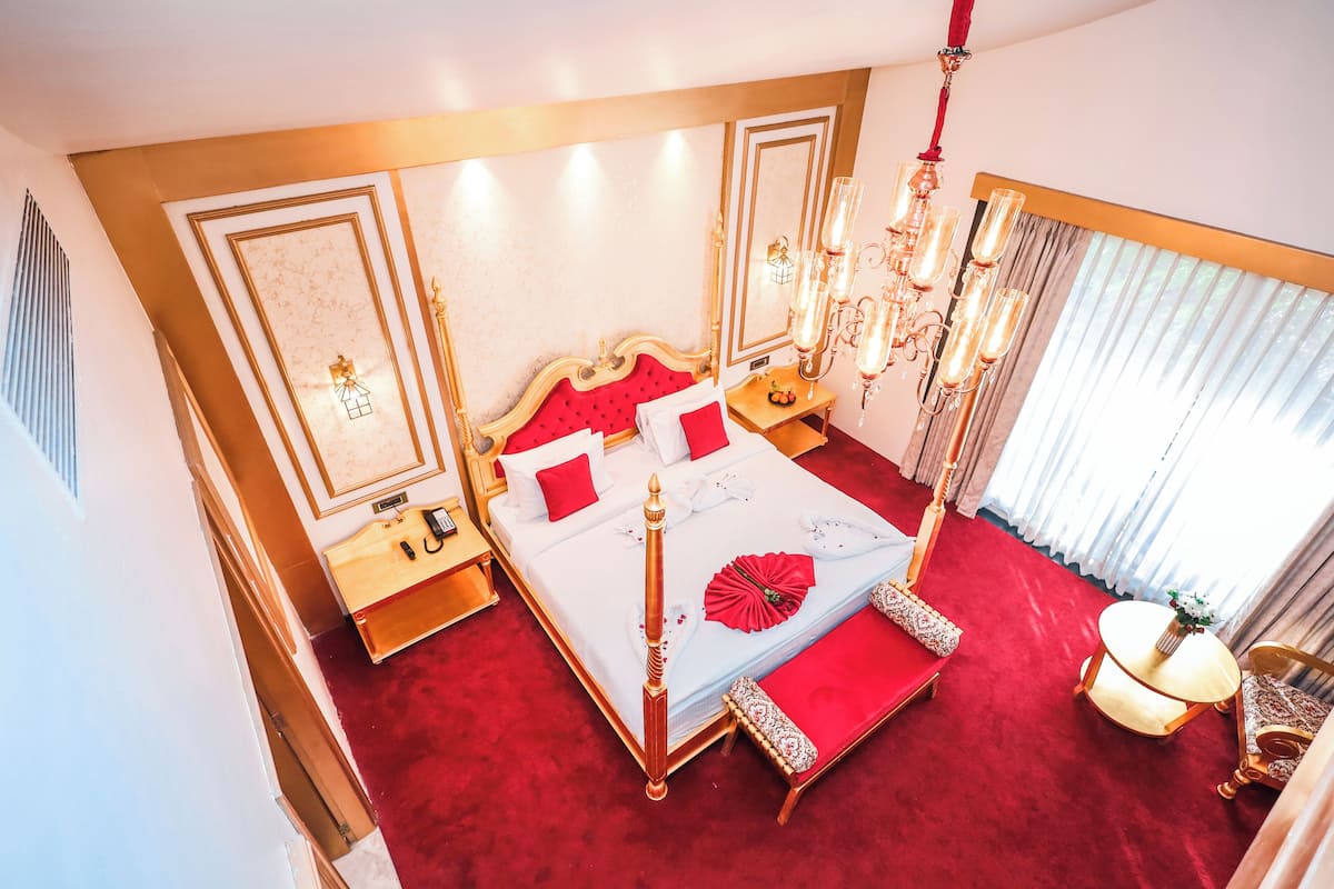 A luxurious bedroom with a red carpet, a large bed with a red headboard, a chandelier, and a bedside table with a lamp.