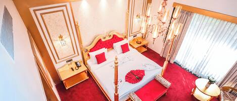A luxurious bedroom with a red carpet, a large bed with a red headboard, a chandelier, and a bedside table with a lamp.