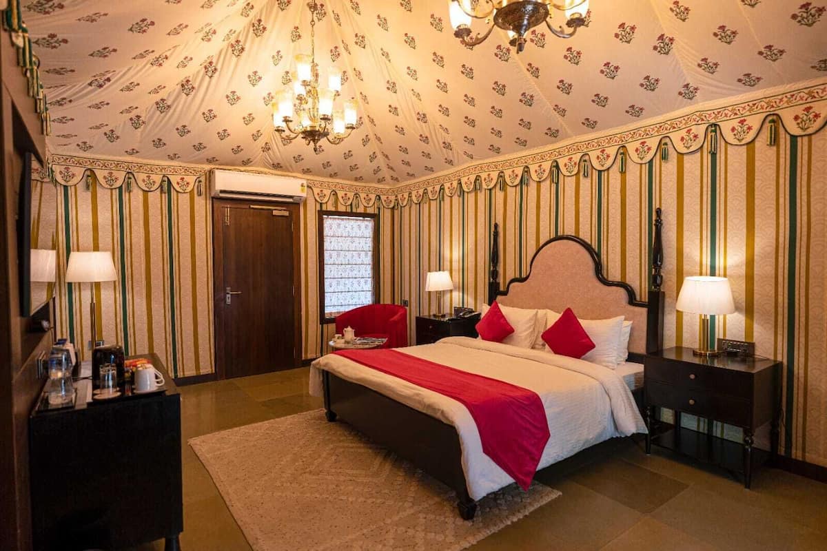 A hotel room with a large bed, two bedside tables with lamps, a red chair, a wooden wardrobe, and a chandelier.