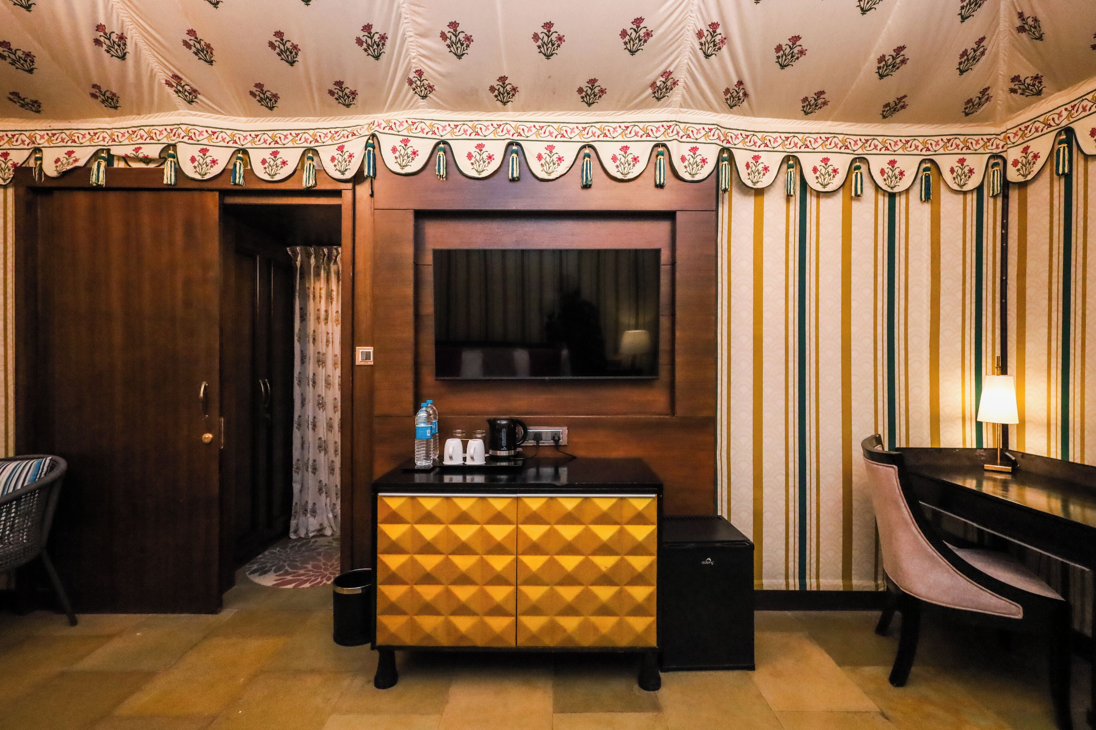 A hotel room with a wooden door, a minibar, a television, a striped wallpaper, and a desk with a lamp.