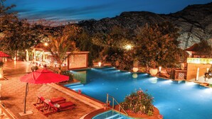 A nighttime swimming pool with red lounge chairs and umbrellas, surrounded by trees and a mountain backdrop. - Estherea Resort and Spa, Jaipur (Bassi)