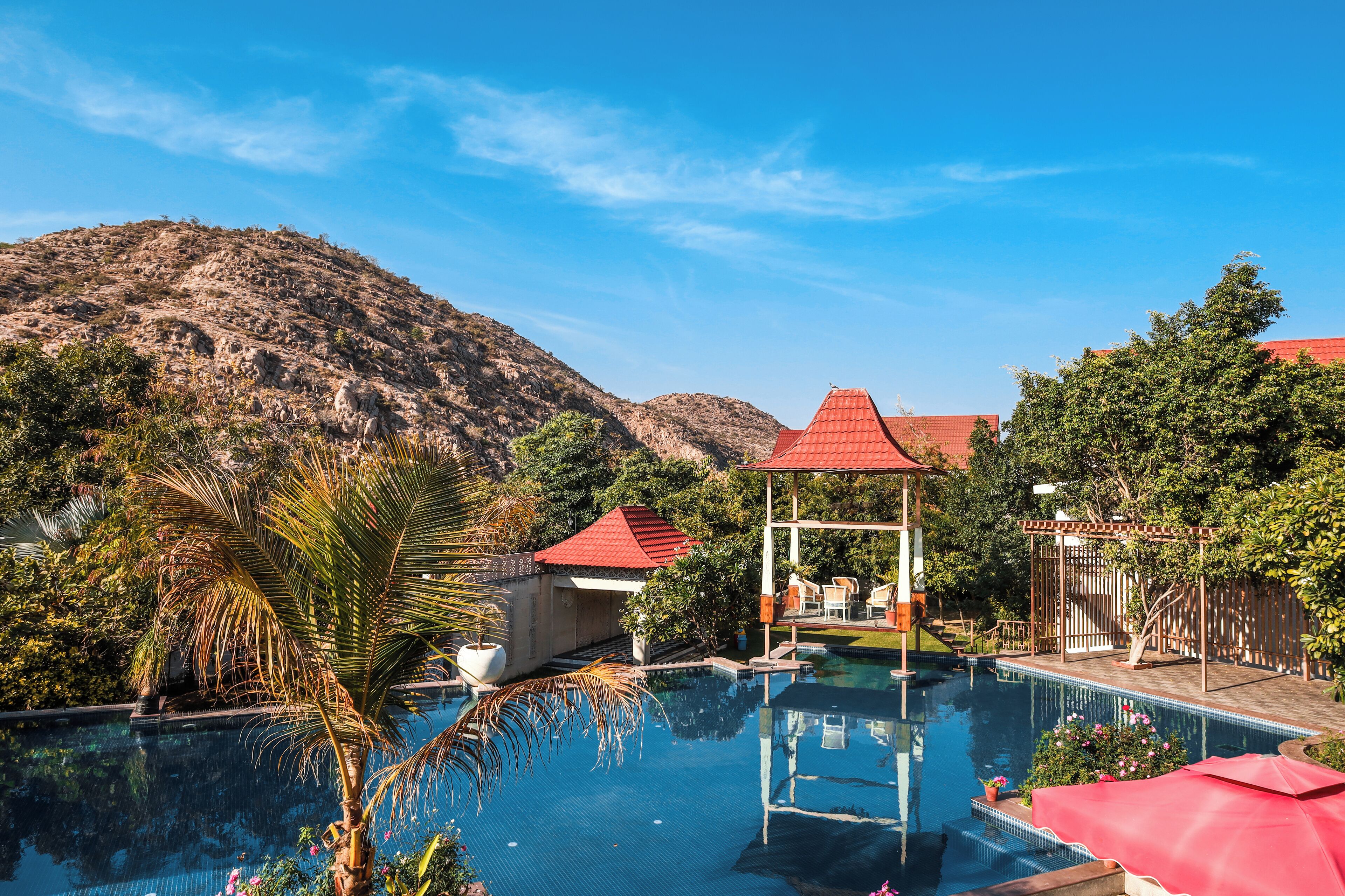 A swimming pool with a red-roofed pavilion, surrounded by lush greenery and a mountainous backdrop.