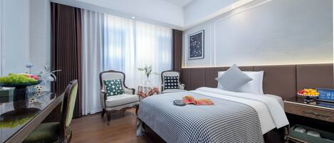 A hotel room with a large bed, a desk with a fruit bowl, a chair, a small table with a vase, and a nightstand with a lamp.