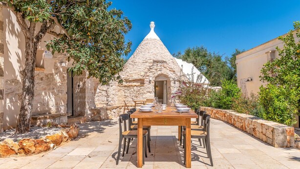A stone building with a conical roof, a wooden table with chairs, and a stone wall with greenery.