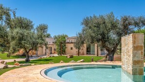 A pool area with stone flooring, surrounded by olive trees and a building with a beige facade.