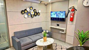 A modern living room with a grey sofa, a round coffee table with a vase of flowers, a flat-screen TV mounted on the wall, a fire extinguisher, and a clock. - HOTEL SKY GOLD (Gandhinagar)