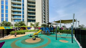 A playground with a slide, swings, and climbing structures, surrounded by a fence and greenery.