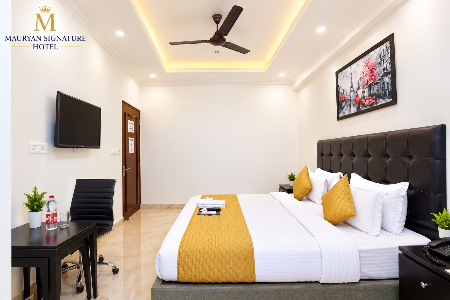 A hotel room with a bed, a desk, a chair, a ceiling fan, a television, and a painting on the wall.
