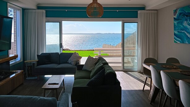 A modern living room with a large sofa, a coffee table, and a dining area with a view of the sea.