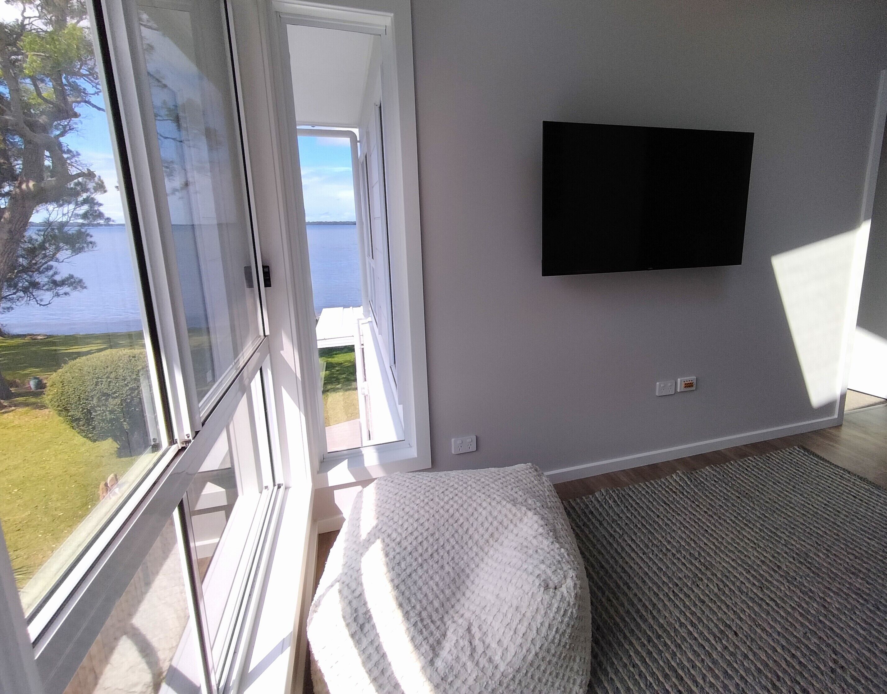 A room with a large window, a flat-screen TV mounted on the wall, and a textured rug on the floor.