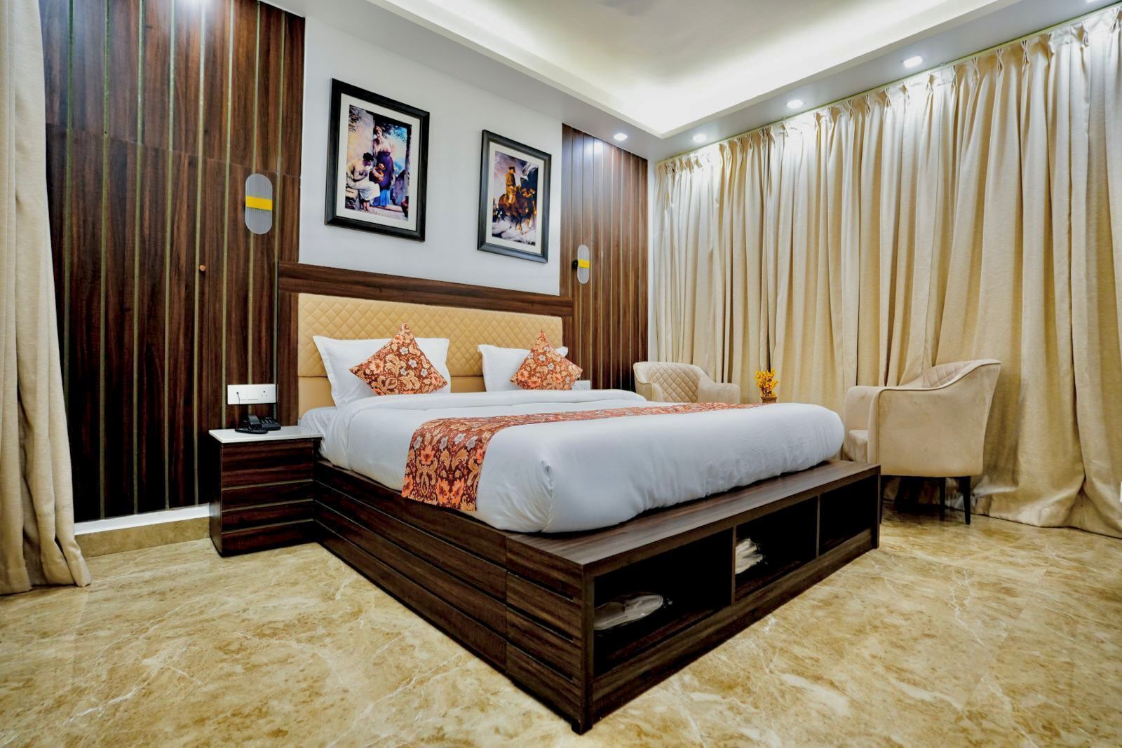 A modern bedroom with a large bed, wooden headboard, and a built-in bench. There are two framed pictures on the wall, a bedside table with a lamp, and a chair.