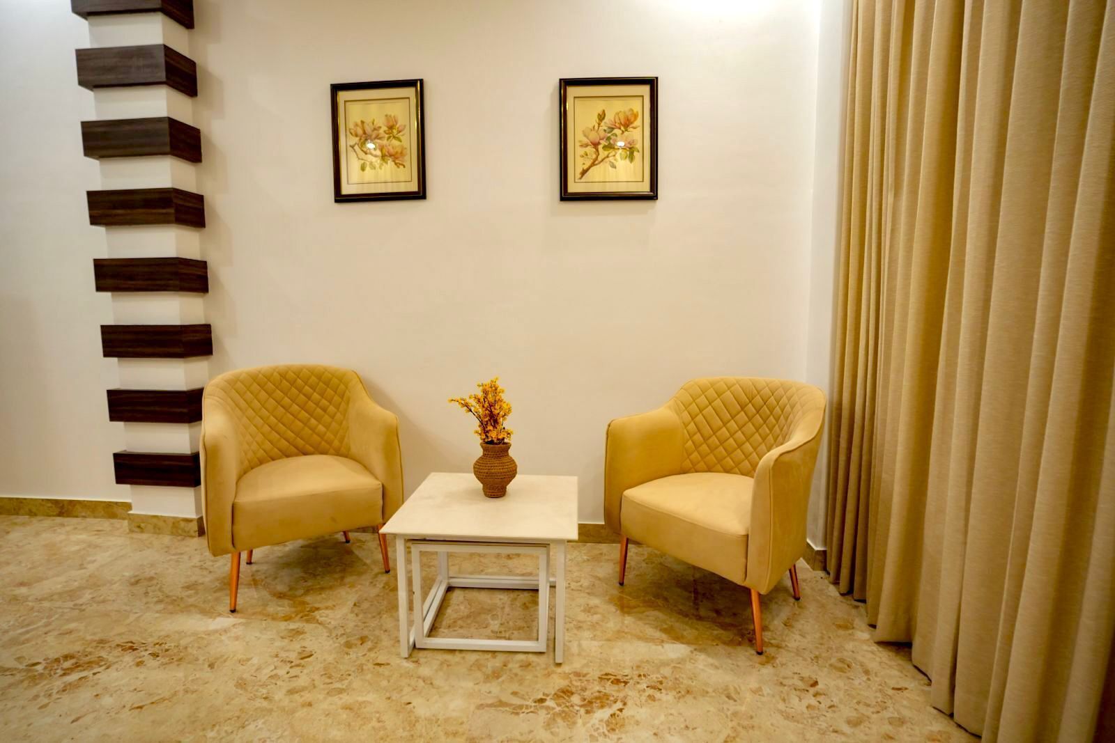 A room with two yellow armchairs, a small white table, and a vase with flowers. There are two framed floral paintings on the wall.