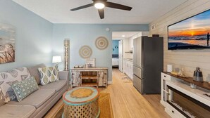 A living room with a sofa, a coffee table, a ceiling fan, and a kitchen area with a refrigerator and a TV. - Beautiful 1BR Condo w/ Ocean View & 2 Heated Pools (North Wildwood)