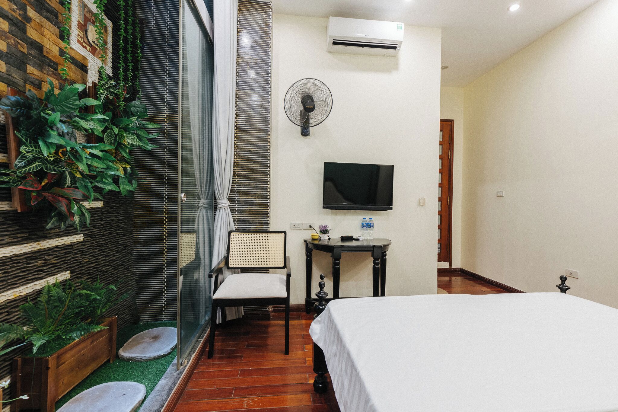 A hotel room with a bed, a TV, a fan, a chair, and a small table.