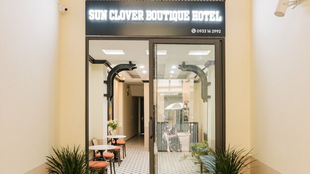 The entrance to Sun Clover Boutique Hotel with a modern design, featuring a glass door, a small outdoor seating area with tables and chairs, and potted plants. - Sun Clover Boutique Hotel (Hanoi)