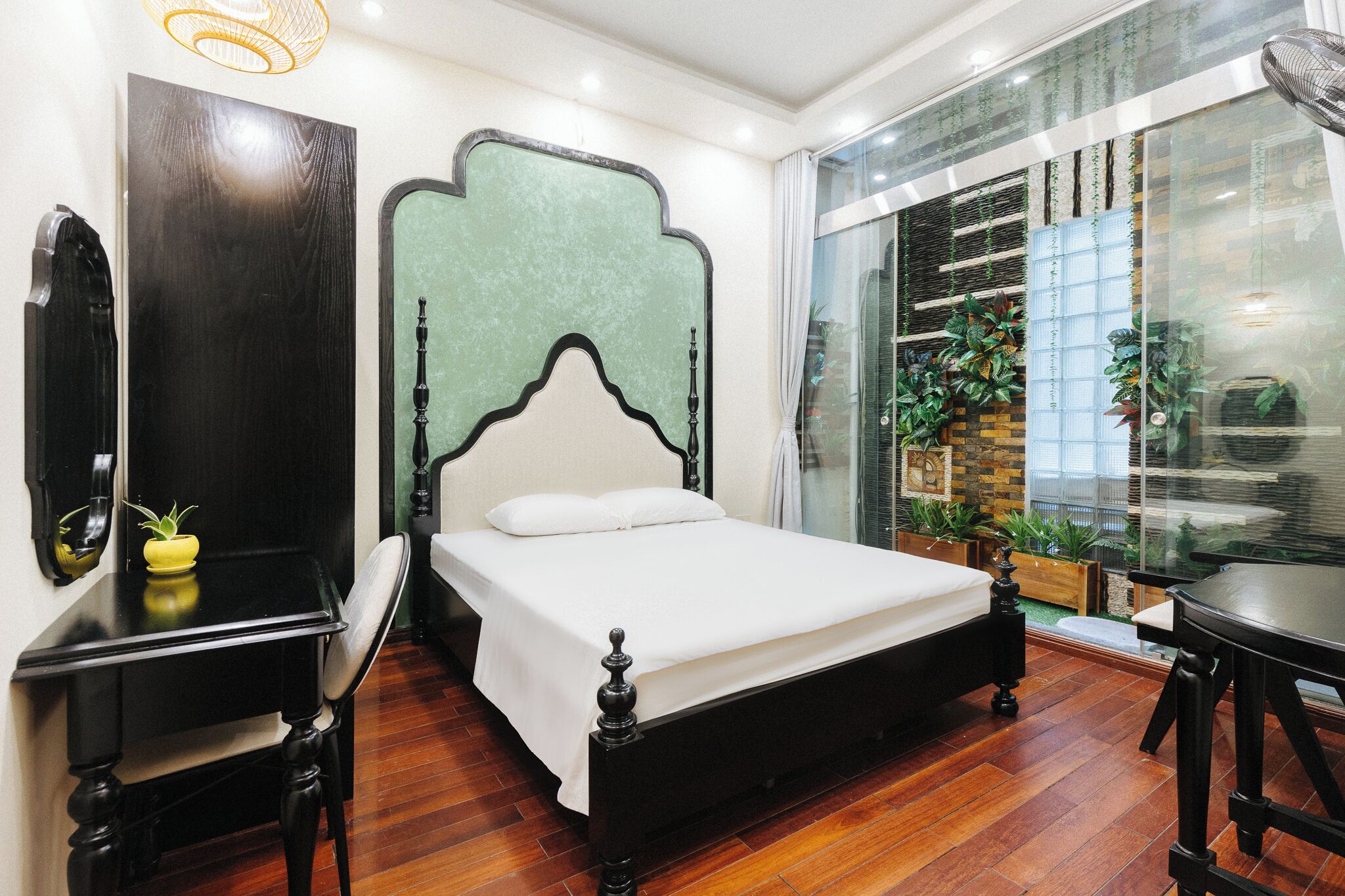 A bedroom with a bed, a nightstand, a chair, a table, and a large window with plants.