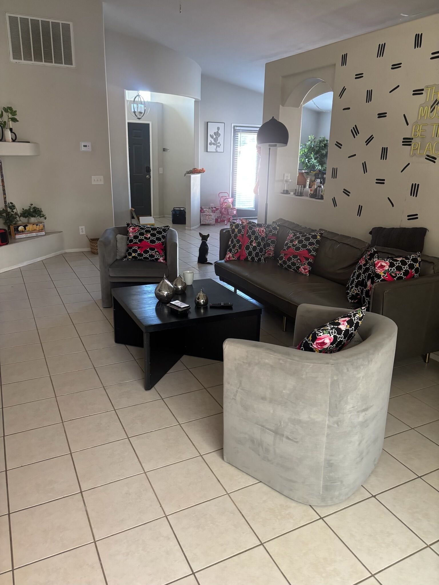 A modern living room with a grey sofa, a black coffee table, and a grey armchair. There is a clock on the wall and a window with blinds.