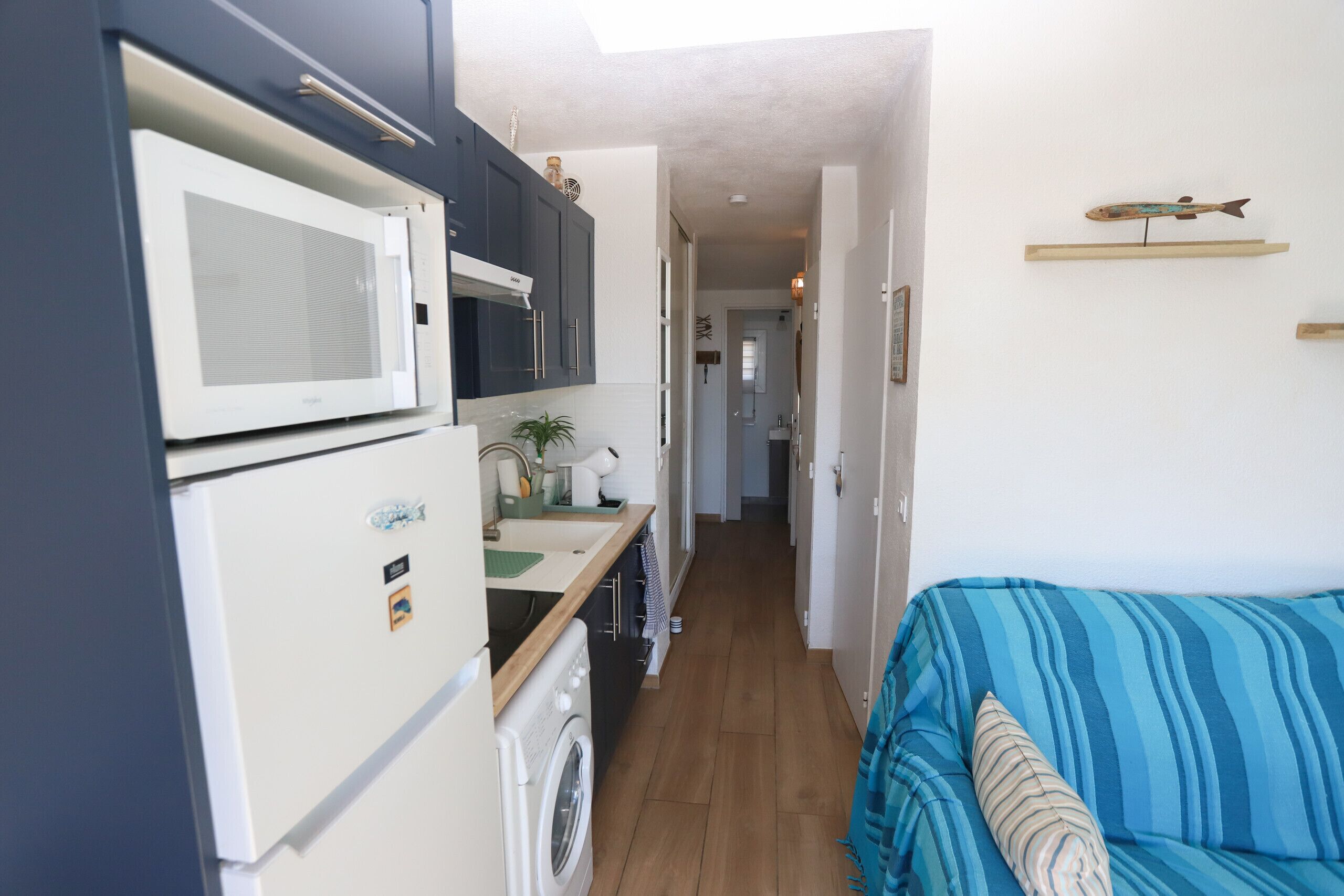 A compact kitchen with a microwave, washing machine, and a small countertop. A blue-striped sofa is visible in the foreground.