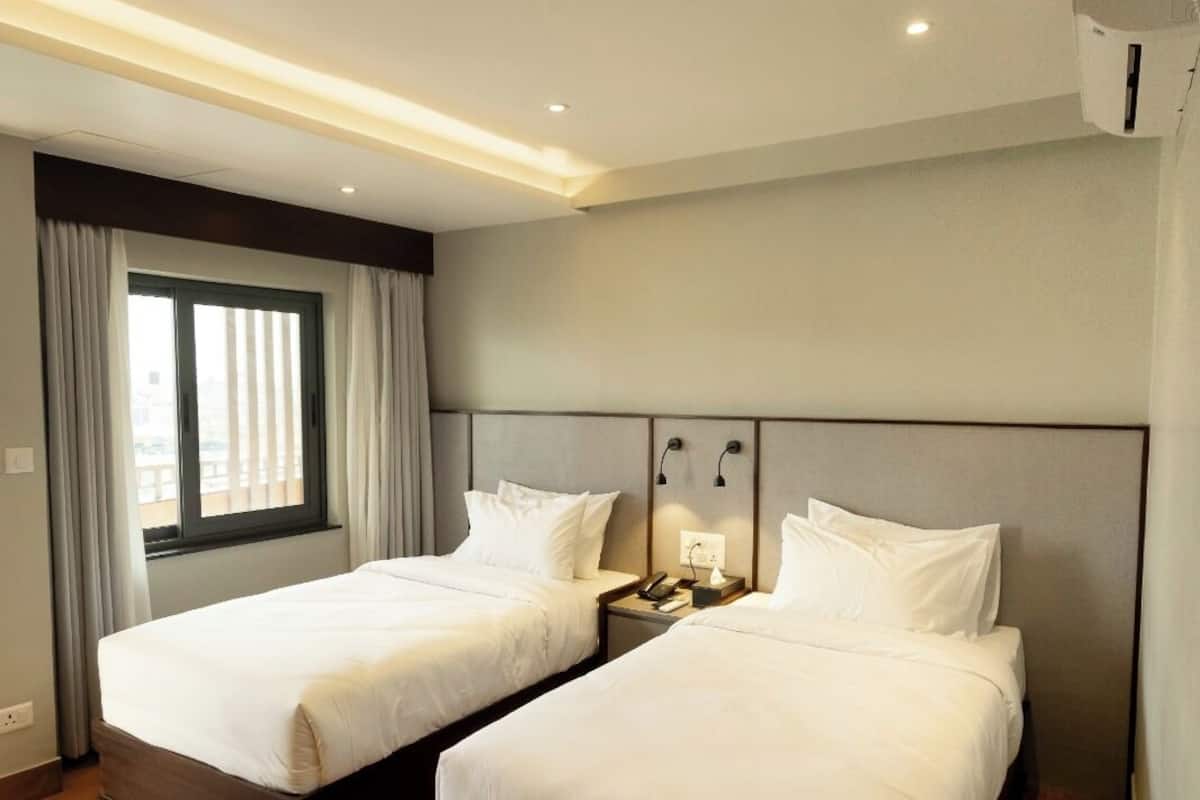 Ein Hotelzimmer mit zwei Betten, Holzboden, einem Fenster mit Vorhängen und einer Klimaanlage.