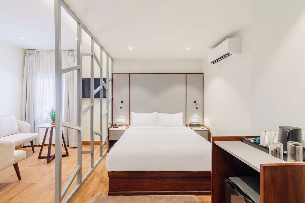 A modern hotel room with a large bed, a desk, a chair, a small table, and a TV.