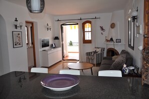 Una sala de estar moderna con un sofá, una mesa de centro y una zona de comedor con mesa y sillas. Se ve una cocina al fondo, con un televisor y una ventana.
