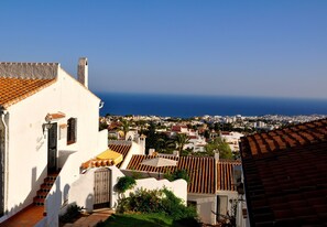 A coastal town with white buildings and red-tiled roofs, a clear blue sky, and the sea in the distance.