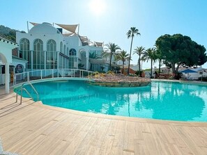 A swimming pool with a wooden deck, surrounded by palm trees and a modern building with arched windows.