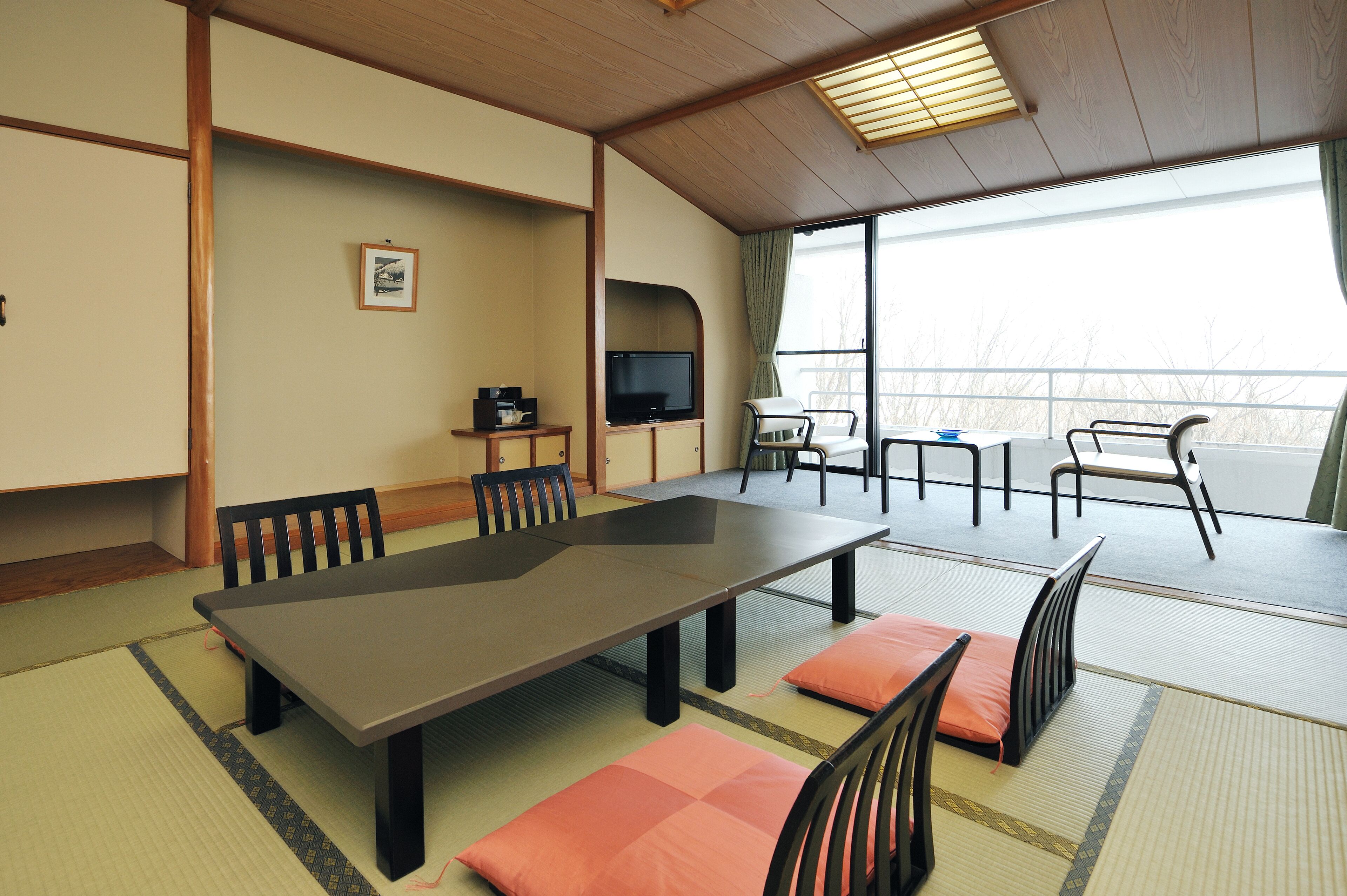 A traditional Japanese-style room with tatami flooring, a low table with cushions, a television, and a view of the outdoors.