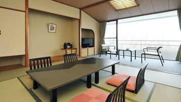 A traditional Japanese-style room with tatami flooring, a low table with cushions, a television, and a view of the outdoors.
