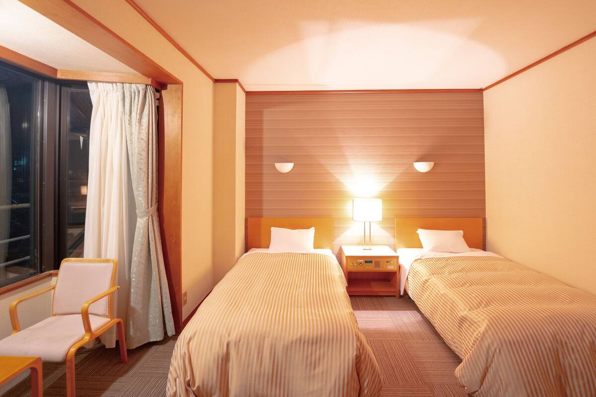 A hotel room with two beds, a nightstand, a lamp, a chair, and a window with curtains.