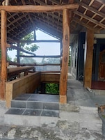 Um espaço ao ar livre coberto, com estrutura de madeira, degraus de pedra e vista para o verde.