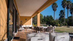 Portico esterno coperto con divano, tavolo da pranzo e sedie, con vista su un prato con palme e cielo sereno.
