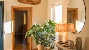 Un'ampia sala d'ingresso con un'abbondante pianta in vaso, un tavolo consolle in legno con oggetti decorativi, uno specchio rotondo e un lampadario intrecciato al soffitto.