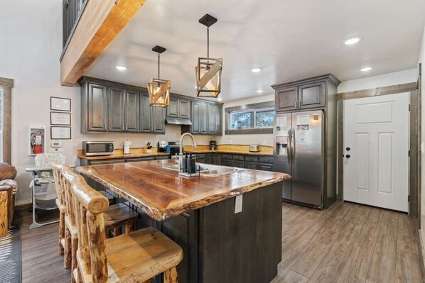 Una cocina con una isla central, taburetes de madera, electrodomésticos de acero inoxidable e iluminación colgante.