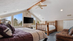 Une chambre avec un grand lit, une tête de lit en bois, un ventilateur de plafond, un téléviseur fixé au mur et une vue sur les arbres à travers de grandes fenêtres.