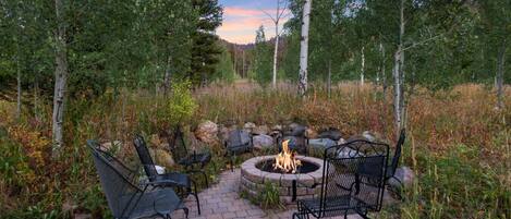 A fire pit surrounded by metal chairs in a forested area with a clear sky.