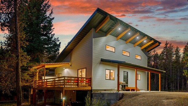 A modern two-story house with a covered deck and a large window overlooking a forested area. - Prism Peaks | Hot Tub, Fire Pit, AC, Yellowstone, ATV, Views! (Island Park)