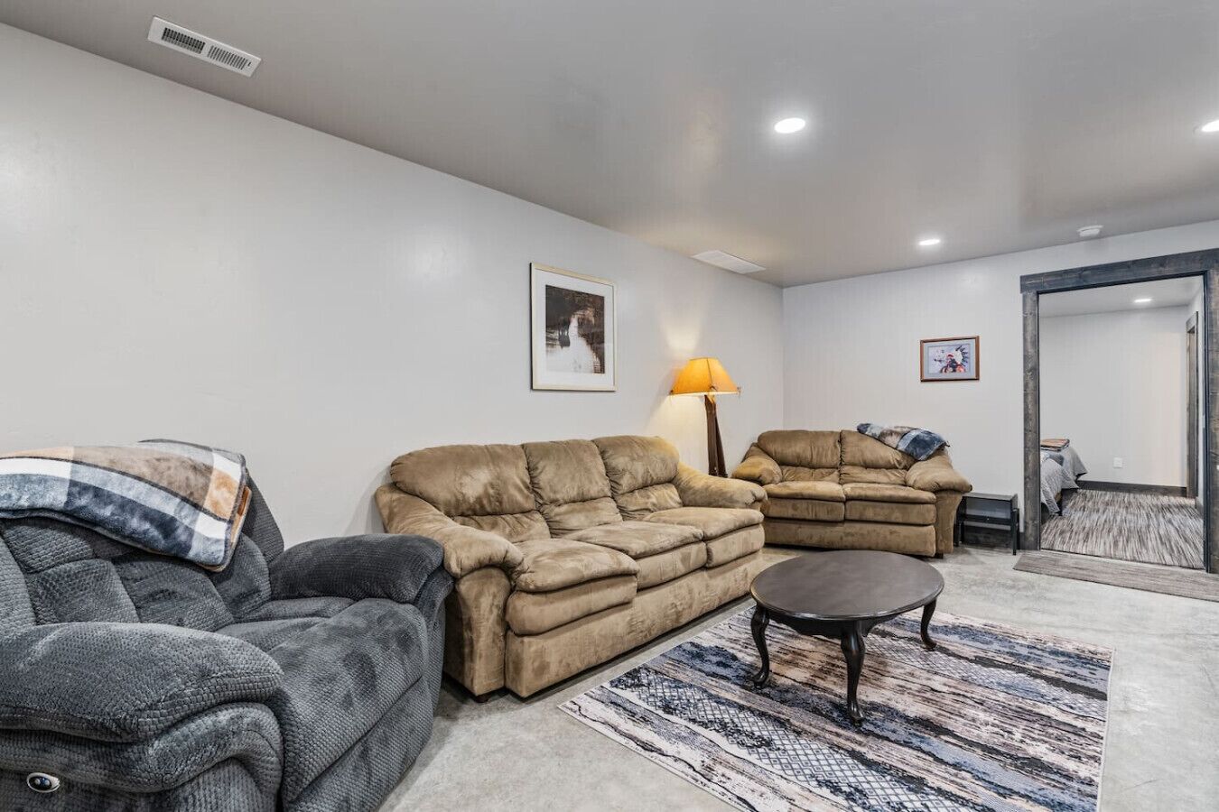 Un salon avec un canapé beige, un fauteuil gris et une petite table ronde. On y trouve une lampe sur une table d’appoint et un tapis sur le sol.