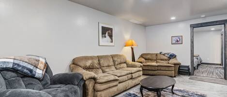 Un salon avec un canapé beige, un fauteuil gris et une petite table ronde. On y trouve une lampe sur une table d’appoint et un tapis sur le sol.