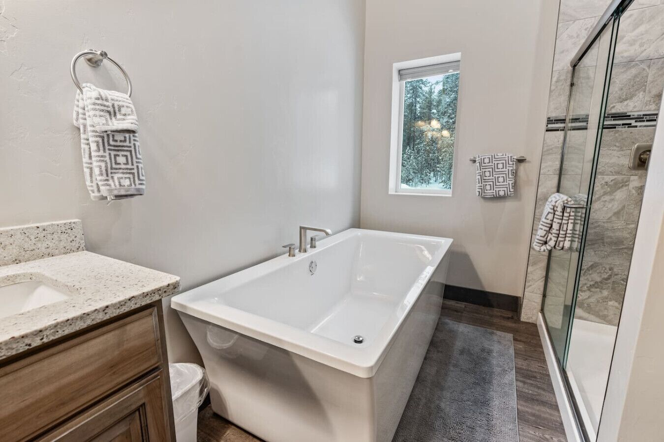 Un baño moderno con una bañera independiente, cabina de ducha con paredes de vidrio y una ventana pequeña con vistas a árboles.