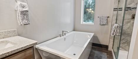 Un baño moderno con una bañera independiente, cabina de ducha con paredes de vidrio y una ventana pequeña con vistas a árboles.
