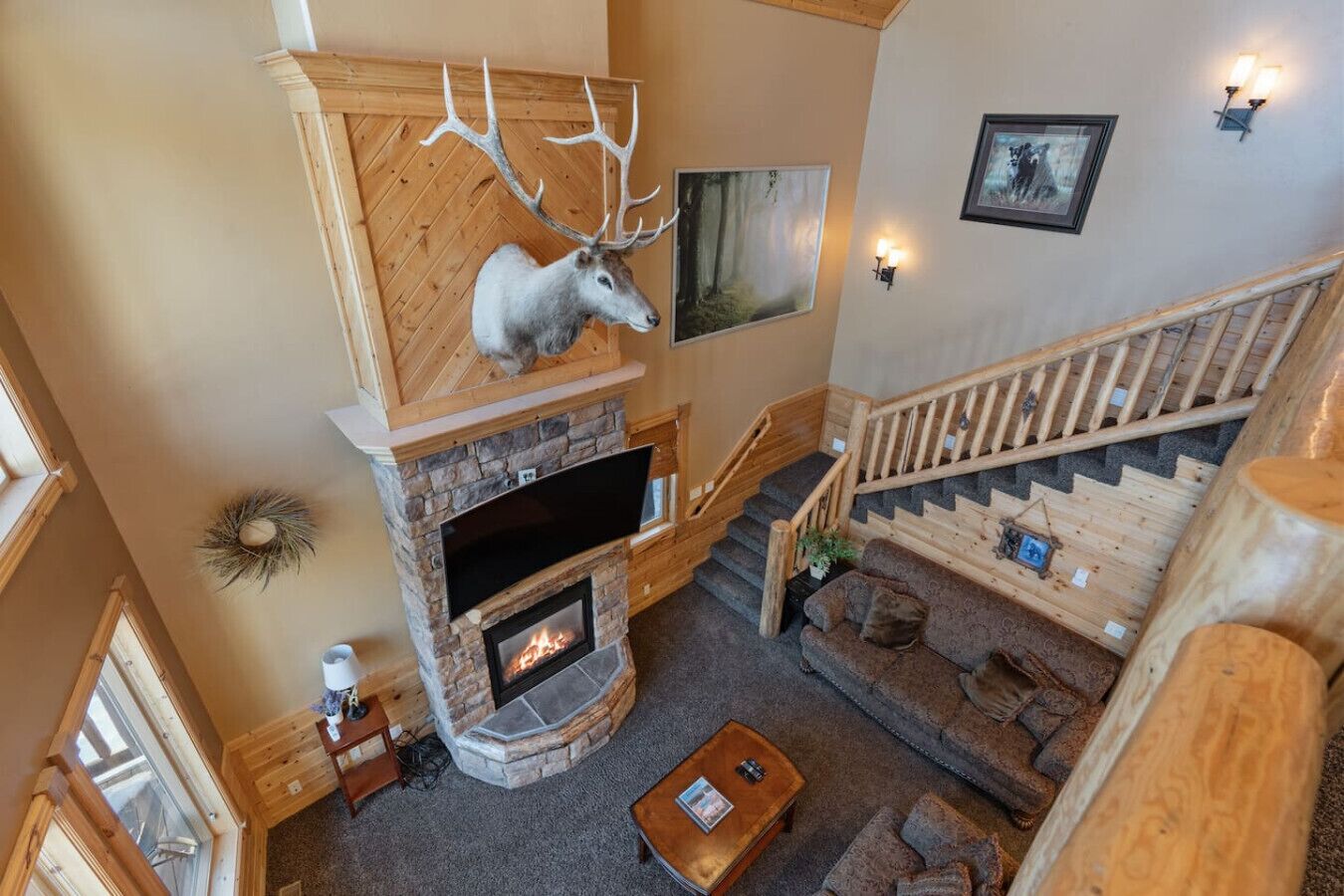 A spacious living area with a stone fireplace, a mounted deer head, and a wooden staircase.