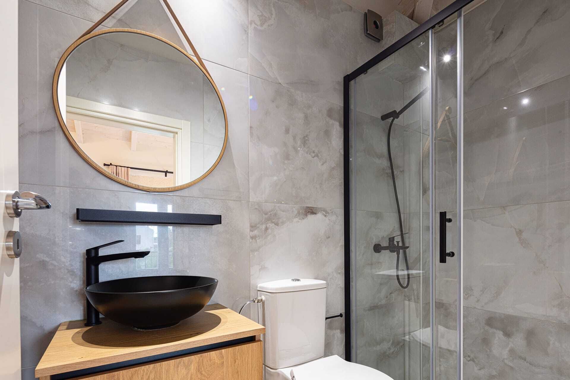 A modern bathroom with a round mirror, black sink, and glass shower enclosure.