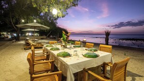 An outdoor dining area with a table set for four, surrounded by wooden chairs, overlooking a beach at sunset.