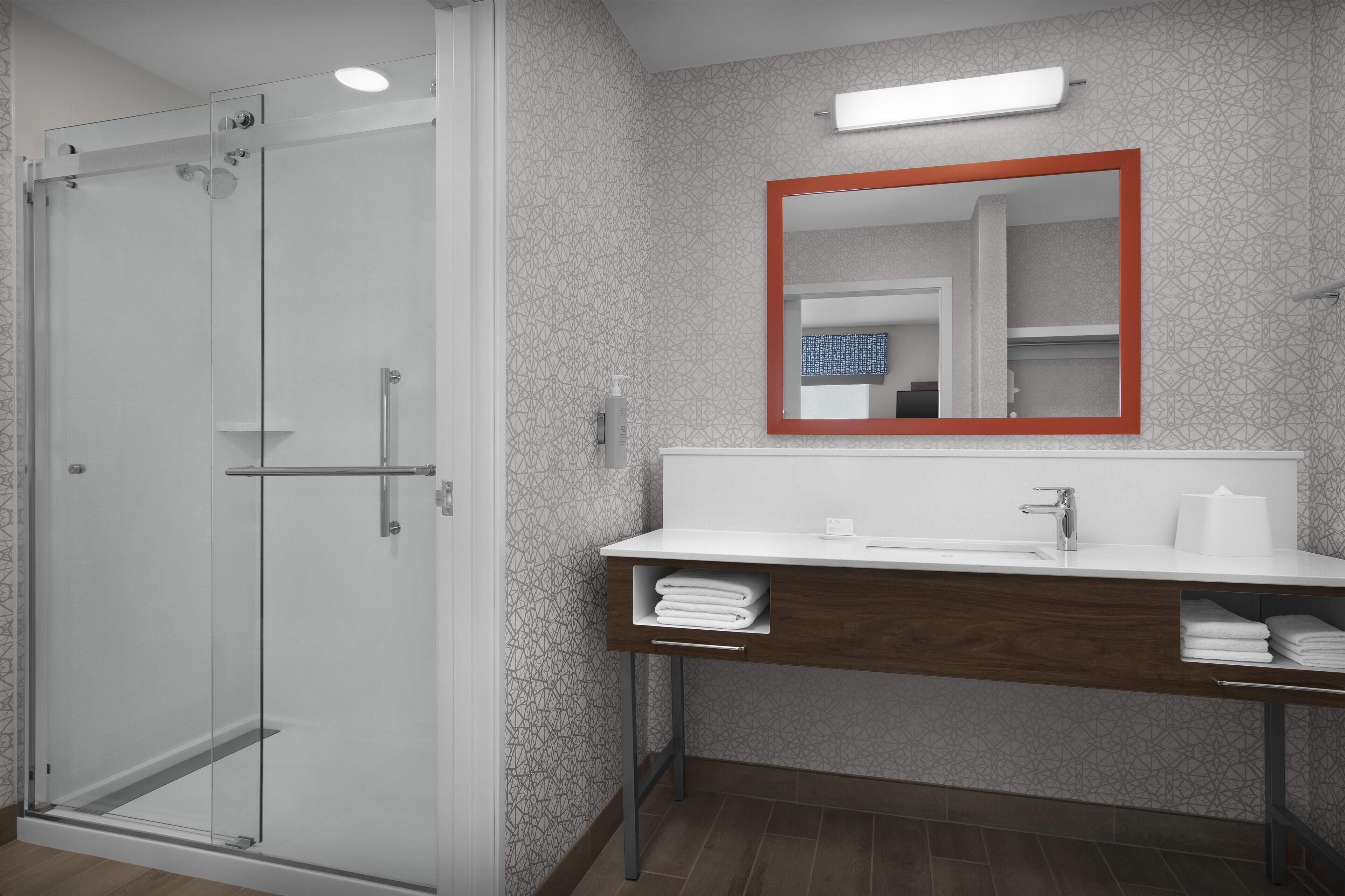 Un baño moderno con cabina de ducha de vidrio, tocador blanco con base de madera y espejo con marco rojo.