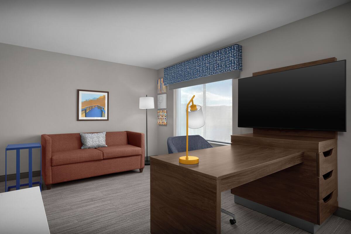 A hotel room with a wooden desk, a flat-screen TV, a sofa, a lamp, and a large window with a view.