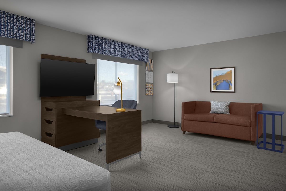 A modern hotel room with a wooden desk, a sofa, a TV, and a floor lamp.