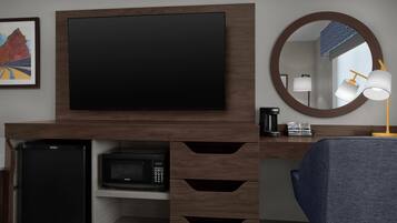 Una habitación moderna con televisión de pantalla plana montada en un mueble de madera, microondas, refrigerador, escritorio con lámpara y una silla.
